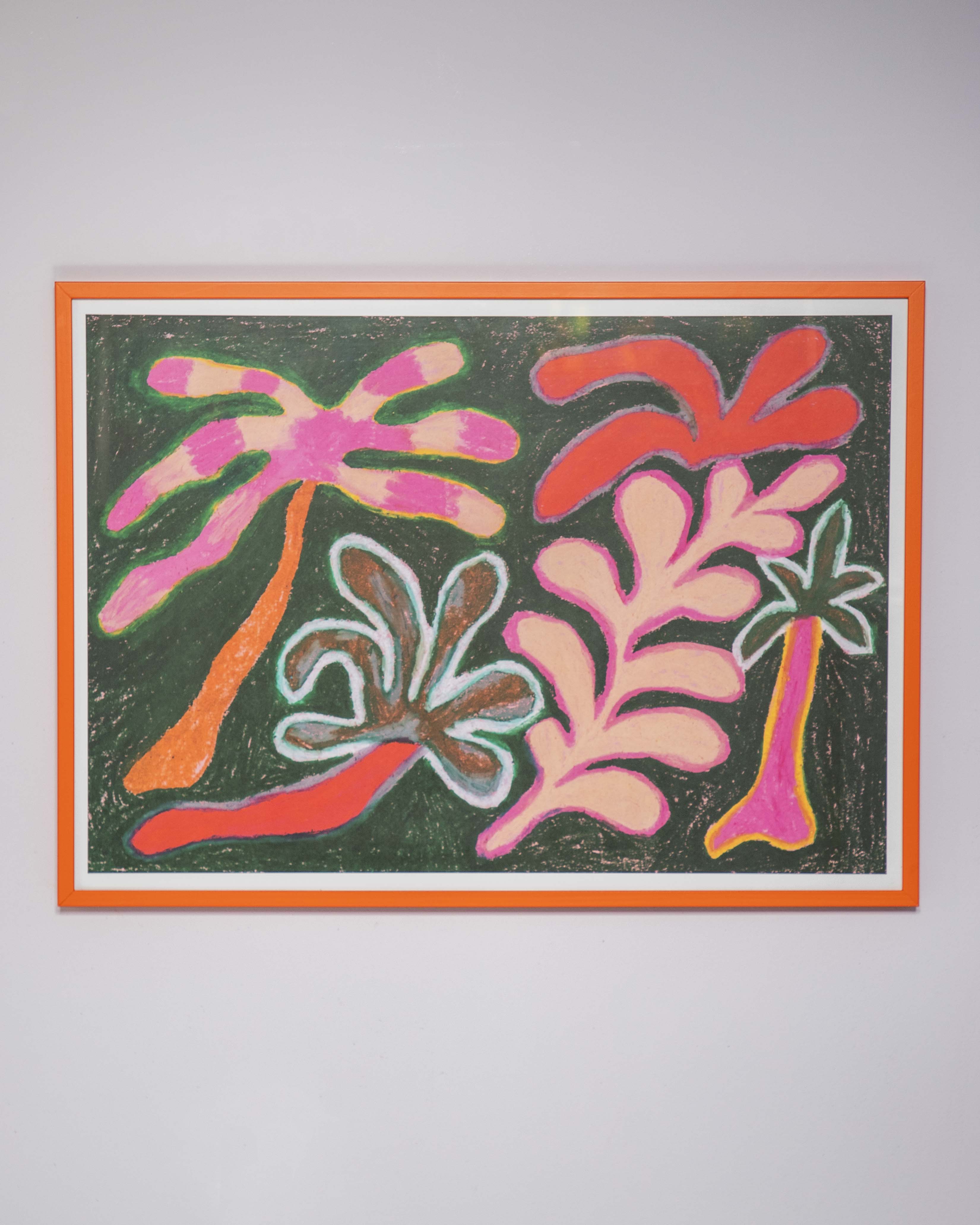 A framed abstract artwork featuring stylized palm-like shapes in pink, coral, orange, and brown on a dark green background, bordered by white and a bright orange frame, mounted on a light gray wall.