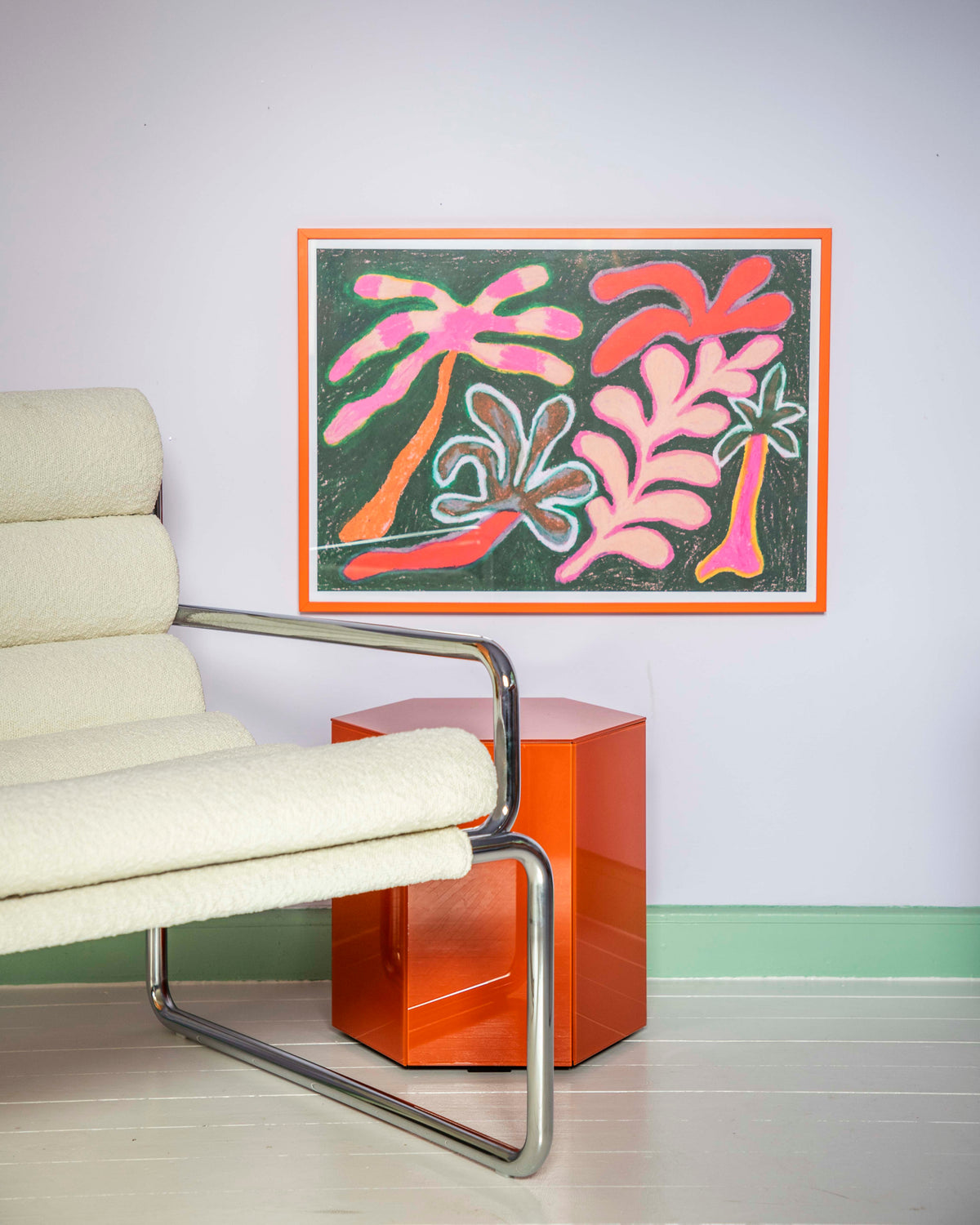 A framed abstract art print featuring pink and orange palm-like shapes on a dark green background, styled in a modern room setting above a low orange side table and next to a cream-colored armchair.